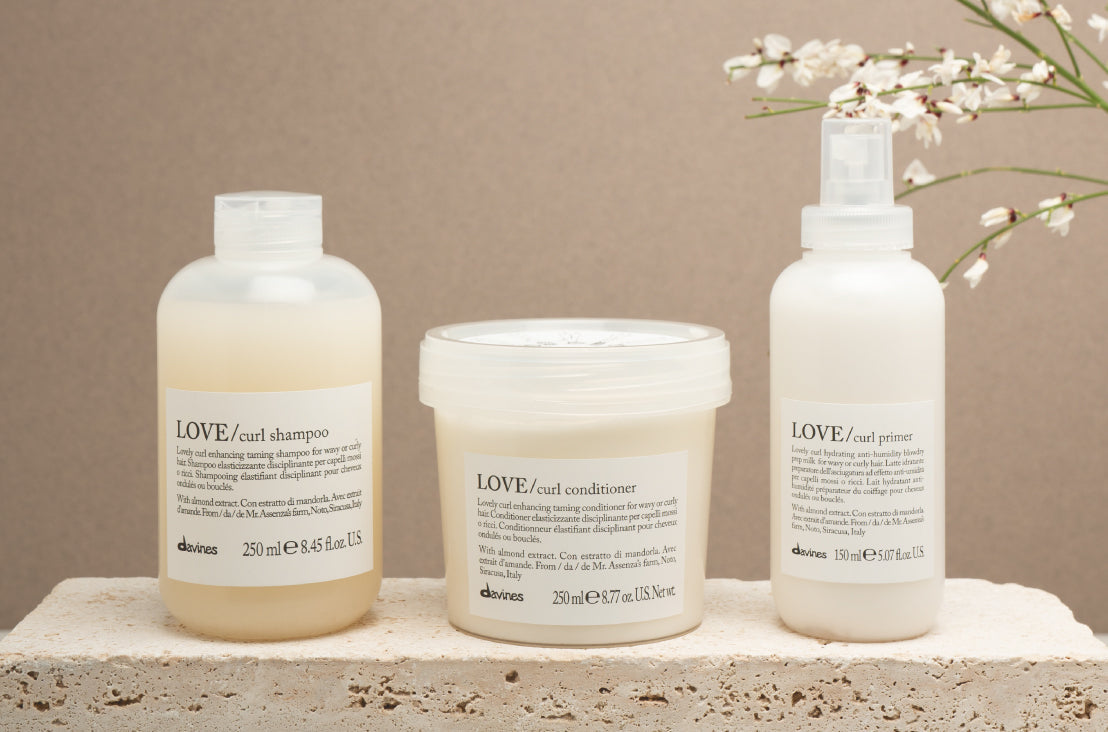 Davines' love curl shampoo, conditioner, and primer displayed on a concrete block with a warm brown background. a small branch of flowers is in the top right corner